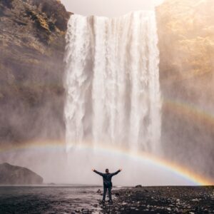 A man raising his hands, looking at the water falls with rainbows