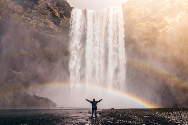 A man raising his hands, looking at the water falls with rainbows